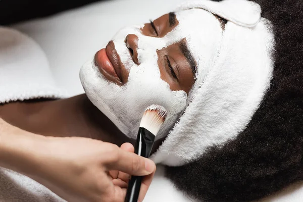 Close up vista da mulher afro-americana positiva com headband deitado perto de terapeuta spa aplicando máscara facial na bochecha no salão de spa — Fotografia de Stock