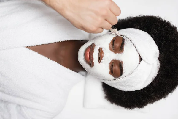 Vue du dessus de femme afro-américaine positive portant un peignoir, couché près d'un thérapeute spa appliquant un masque facial sur la joue dans un salon de spa — Photo de stock