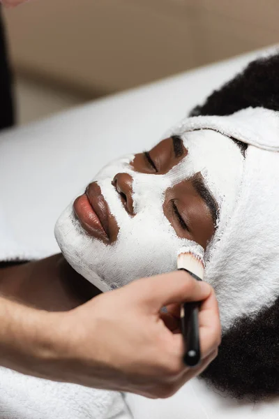 Vista da vicino della donna afro-americana con fascia distesa vicino al terapeuta spa che applica la maschera facciale sulla guancia nel salone spa — Foto stock