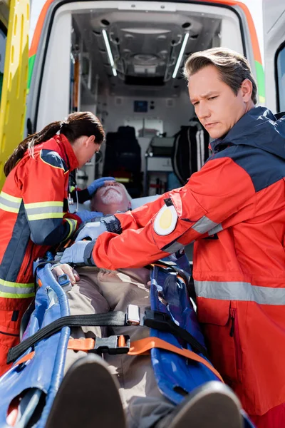 Selective focus of paramedics standing near elderly patient on stretcher and ambulance car — Stock Photo