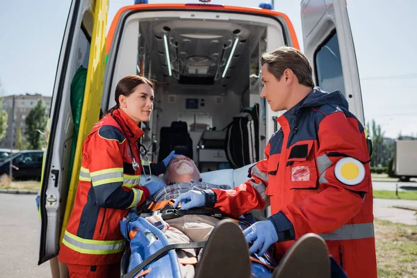 Enfoque selectivo de los paramédicos en guantes de látex de pie cerca del paciente en camilla y coche ambulancia al aire libre - foto de stock
