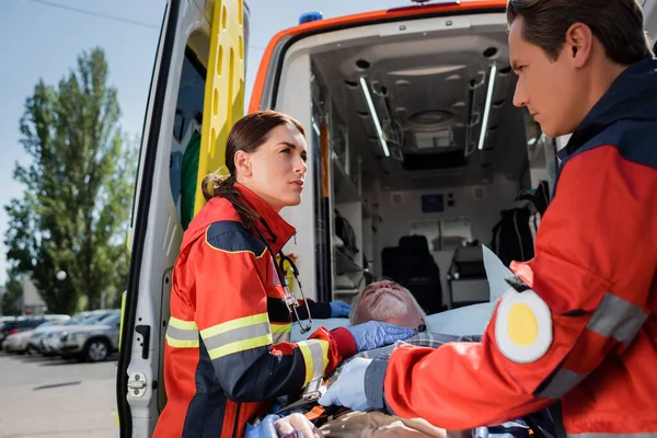 Concentration sélective des ambulanciers paramédicaux en uniforme debout près du patient âgé sur civière et ambulance auto sur la rue urbaine — Photo de stock