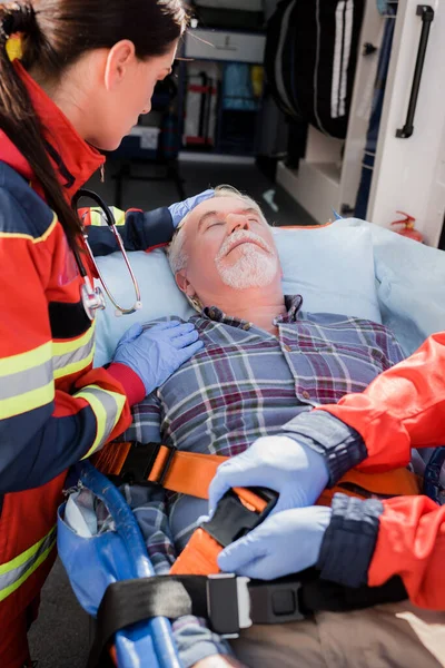 Selective focus of paramedic touching patient while colleague locking belts of stretcher and ambulance car — Stock Photo