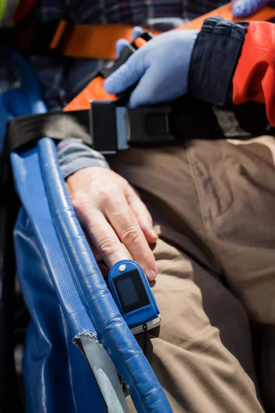 Cropped view of paramedic in latex gloves locking belt of stretcher near patient with heart rate monitor — Stock Photo