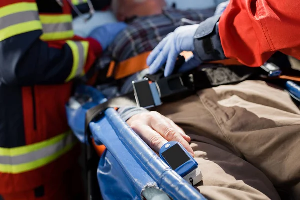 Vista recortada del paciente con monitor de frecuencia cardíaca acostado en camilla cerca de paramédicos - foto de stock