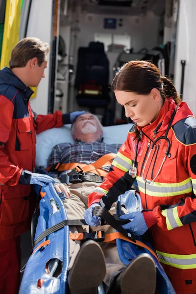 Selective focus of paramedic with stethoscope fastening belt of stretcher near patient and colleague — Stock Photo