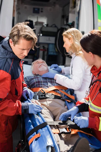Selective focus of paramedics and doctor standing near senior patient on stretcher and ambulance car — Stock Photo
