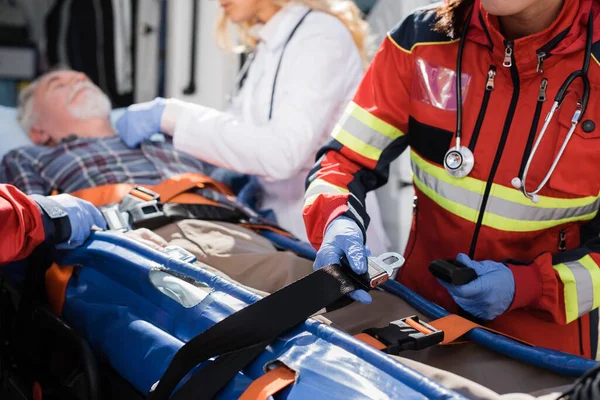 Selective focus of paramedic holding belts of stretcher near patient and colleagues outdoors — Stock Photo