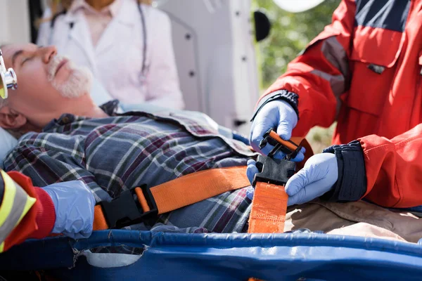 Selective focus of paramedic locking belts of stretcher near elderly patient and colleagues outdoors — Stock Photo