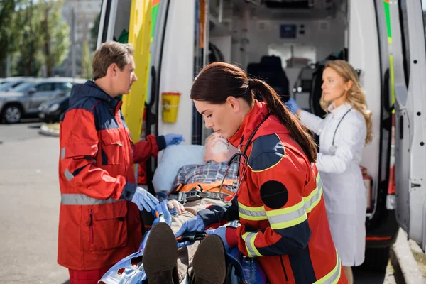 Selective focus of paramedic fastening belts near patient on stretcher and colleagues on urban street — Stock Photo