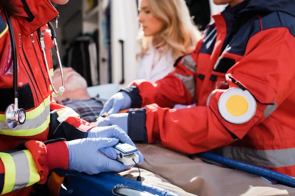 Focus selettivo del cardiofrequenzimetro paramedico vicino al paziente su barella e colleghi all'aperto — Foto stock