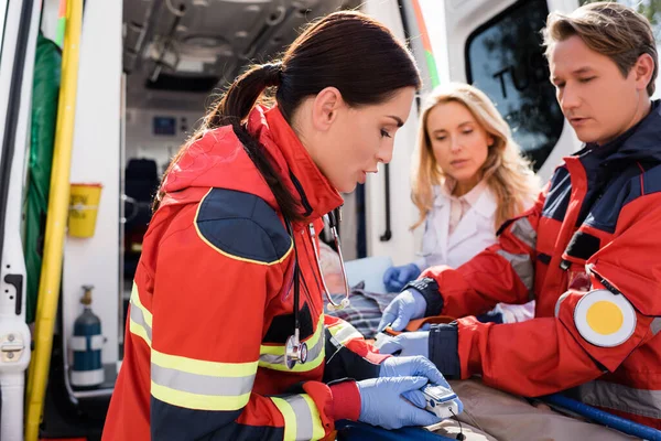 Concentration sélective de l'ambulancier dans les gants en latex tenant le moniteur de fréquence cardiaque près du patient âgé sur civière et ses collègues à l'extérieur — Photo de stock