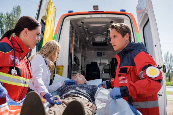 Selective focus of paramedics and doctor standing near senior man on stretcher and ambulance car outdoors — Stock Photo