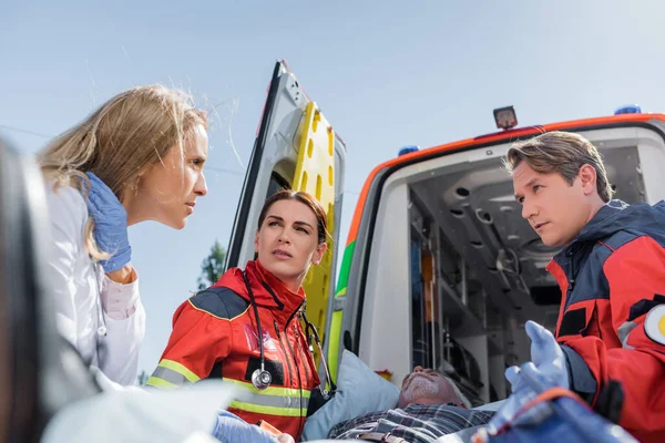 Selective focus of doctor looking at paramedic near patient on stretcher and ambulance car on urban street — Stock Photo