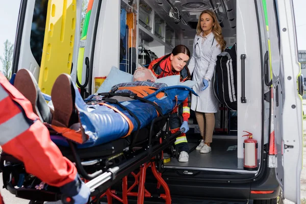 Выборочный фокус врача, стоящего рядом с парамедиками, держащего носилки с пациентом в машине скорой помощи — стоковое фото