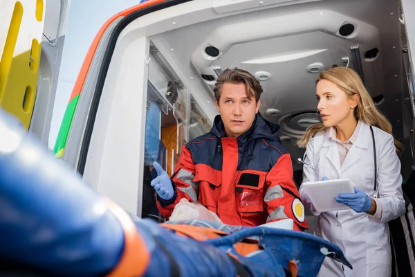 Selective focus of paramedic pointing at senior patient on stretcher near doctor with digital tablet in ambulance car — Stock Photo