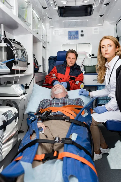 Выборочный фокус врача, смотрящего в камеру рядом со старшим пациентом и парамедиком в машине скорой помощи — стоковое фото