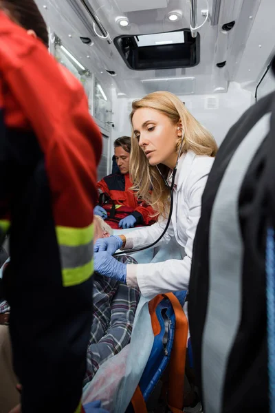 Enfoque selectivo del médico que examina al paciente con estetoscopio cerca de los paramédicos en el coche de ambulancia - foto de stock