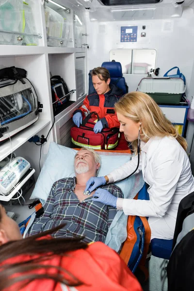 Concentration sélective du médecin en gants de latex examinant le patient âgé avec stéthoscope près des ambulanciers en uniforme dans la voiture d'ambulance — Photo de stock