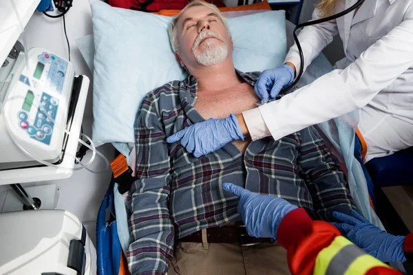 Selective focus of doctor with stethoscope and paramedic pointing with fingers near senior patient in ambulance car — Stock Photo