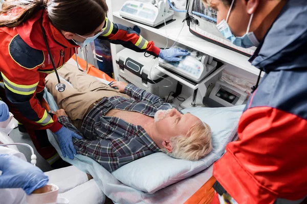 Focus selettivo dei paramedici in maschere mediche e medico in piedi vicino al paziente anziano in auto ambulanza — Foto stock