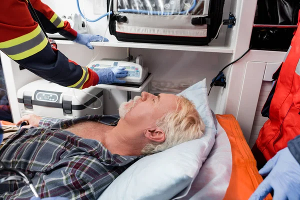 Selective focus of elderly patient lying near paramedics in latex gloves in ambulance car — Stock Photo