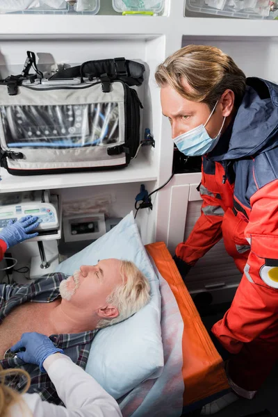 Concentration sélective de l'ambulancier dans le masque médical debout près du médecin examinant le patient âgé avec stéthoscope dans la voiture d'ambulance — Photo de stock