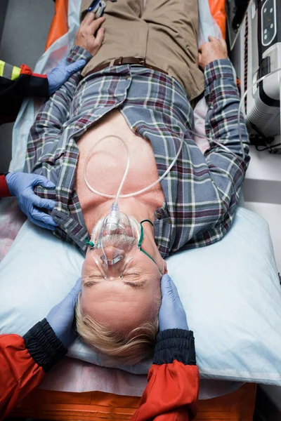 Top view of patient in oxygen mask lying near paramedics in latex gloves in ambulance car — Stock Photo