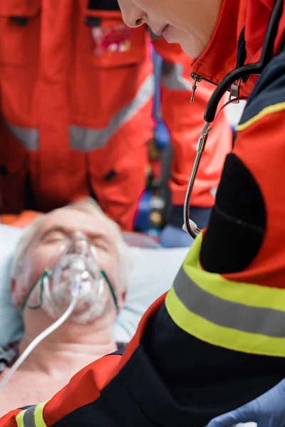 Enfoque selectivo de paramédico en uniforme de pie cerca del anciano en máscara de oxígeno durante los primeros auxilios - foto de stock