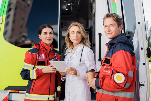 Medico con tablet digitale guardando la fotocamera vicino ai paramedici e auto ambulanza — Foto stock