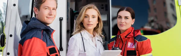 Imagen panorámica del médico con tableta digital mirando a la cámara cerca de paramédicos y ambulancia auto — Stock Photo
