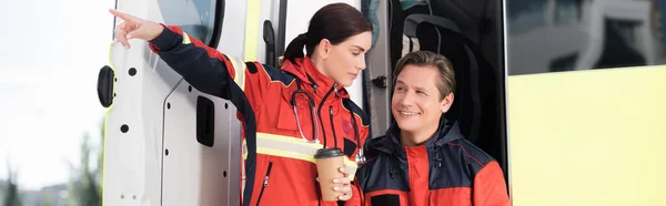 Horizontal image of paramedic looking away near colleague with paper cup pointing with finger and ambulance car — Stock Photo