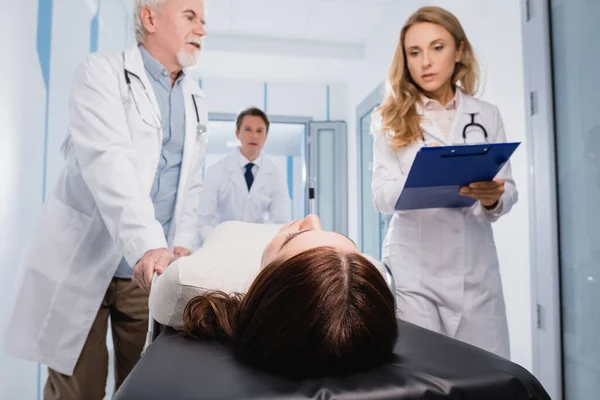 Selective focus of doctors with clipboard walking near patient on stretcher in clinic — Stock Photo