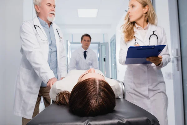 Selective focus of sick woman lying on stretcher near doctors with clipboard in clinic — Stock Photo