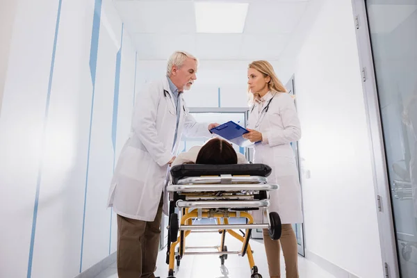 Doctor holding clipboard near senior colleague and sick patient on stretcher in clinic — Stock Photo