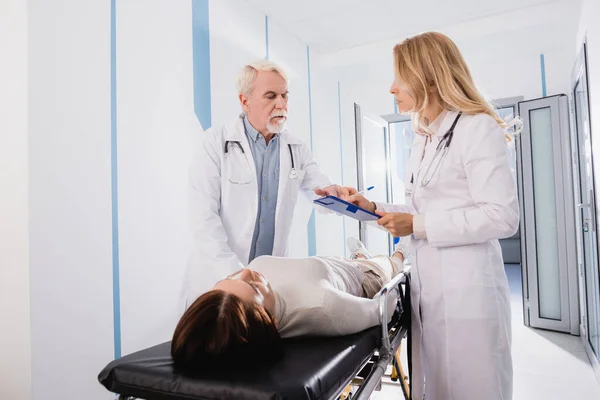 Selective focus of doctor writing on clipboard near senior colleague and diseased patient on stretcher in clinic — Stock Photo