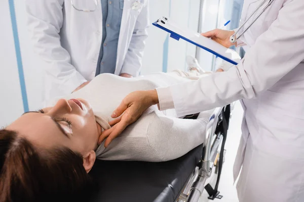 Concentration sélective du médecin avec presse-papiers vérifier le pouls de la femme avec les yeux fermés sur civière en clinique — Photo de stock