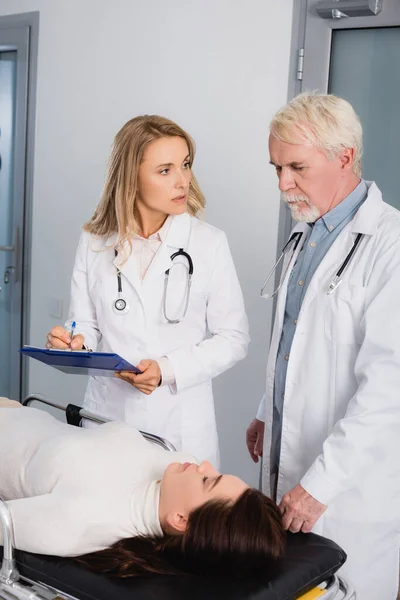 Doctor escribiendo en portapapeles mientras mira a su colega cerca del paciente enfermo en la clínica - foto de stock