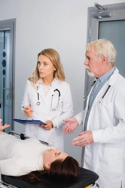 Selective focus of doctors with clipboard looking at colleague near patient in clinic — Stock Photo