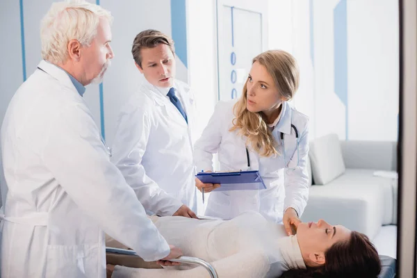 Selective focus of doctor with clipboard checking pulse of patient near colleagues in clinic — Stock Photo