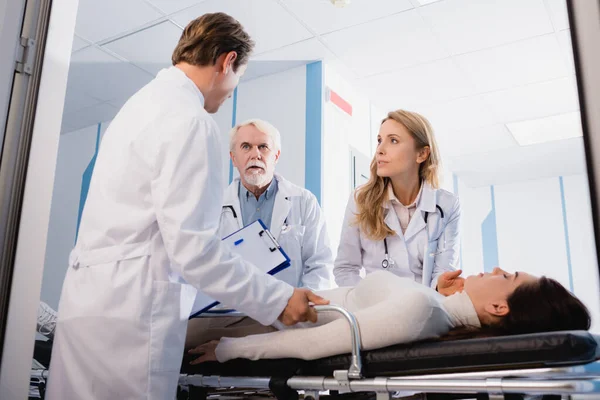 Selective focus of doctors looking at colleague with clipboard near diseased patient with closed eyes on stretcher in corridor of clinic — Stock Photo