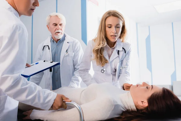 Selective focus of doctors with clipboard looking at woman with closed eyes on stretcher in corridor of clinic — Stock Photo