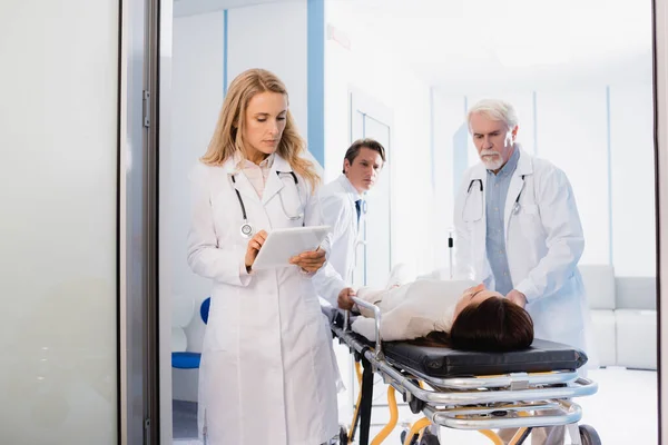 Selective focus of doctor using digital tablet near colleagues and patient on stretcher in clinic — Stock Photo