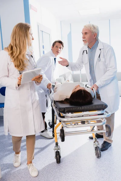 Médico sênior conversando com colega com tablet digital perto do paciente doente na maca na clínica — Fotografia de Stock