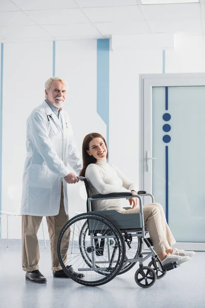 Сеньор врач, стоящий рядом с пациентом в инвалидной коляске в клинике — стоковое фото
