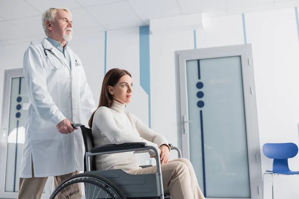 Selective focus of woman sitting in wheelchair near senior doctor in white coat in clinic — Stock Photo