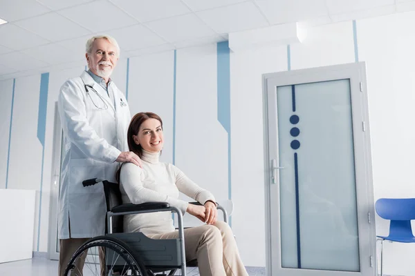 Пацієнт у інвалідному візку дивиться на камеру біля літнього лікаря в клініці — стокове фото