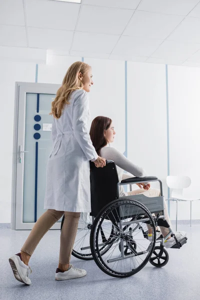 Doctor in white coat walking near disabled patient in wheelchair in clinic — Stock Photo