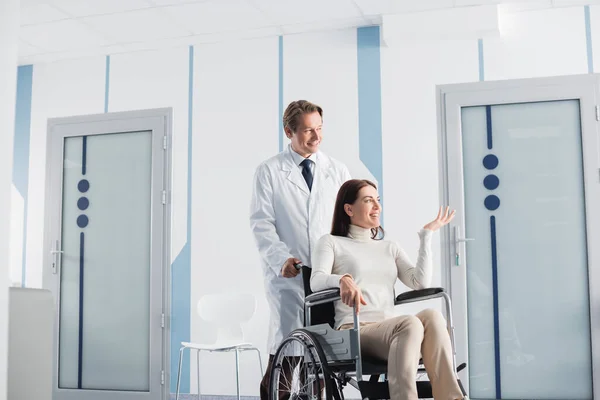 Brunette woman in wheelchair pointing with hand while sitting in wheelchair near doctor in hospital — Stock Photo