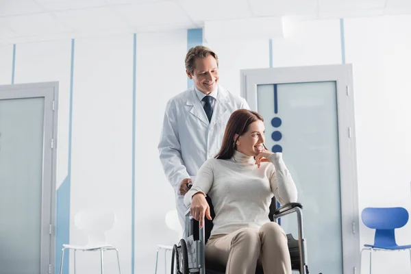 Médico mirando al paciente en silla de ruedas en la clínica - foto de stock